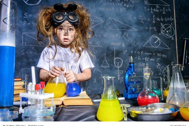 Ein Kind mit Schutzbrille und zerzausten Haaren steht vor Laborgeräten mit bunten Flüssigkeiten, im Hintergrund eine Tafel mit Formeln und Zeichnungen.