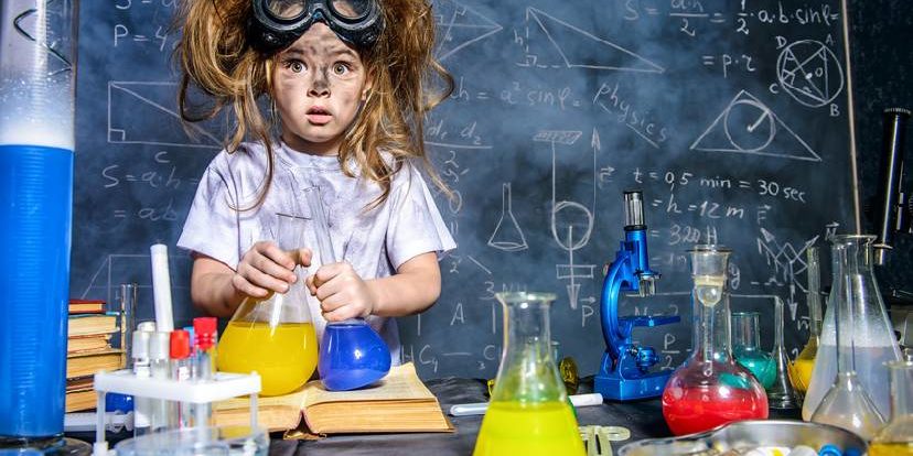 Ein Kind mit Schutzbrille und zerzausten Haaren steht vor Laborgeräten mit bunten Flüssigkeiten, im Hintergrund eine Tafel mit Formeln und Zeichnungen.