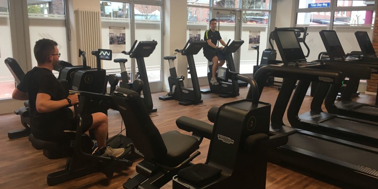 Mehrere Menschen trainieren auf Fitnessgeräten in einem hellen Fitnessstudio mit großen Fenstern und Holzboden.