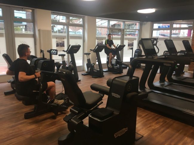 Mehrere Menschen trainieren auf Fitnessgeräten in einem hellen Fitnessstudio mit großen Fenstern und Holzboden.
