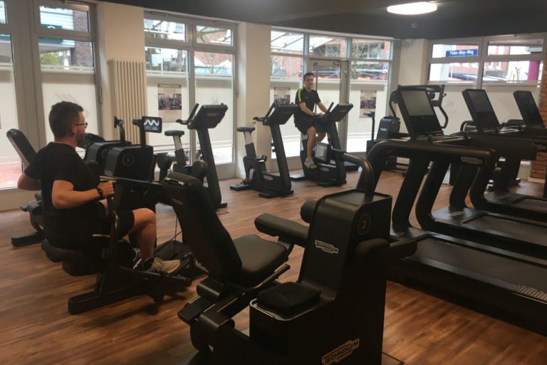 Mehrere Menschen trainieren auf Fitnessgeräten in einem hellen Fitnessstudio mit großen Fenstern und Holzboden.