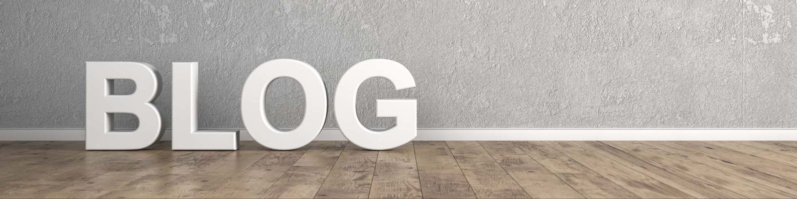 Große weiße Buchstaben "BLOG" stehen auf einem Holzfußboden vor einer grauen Wand.