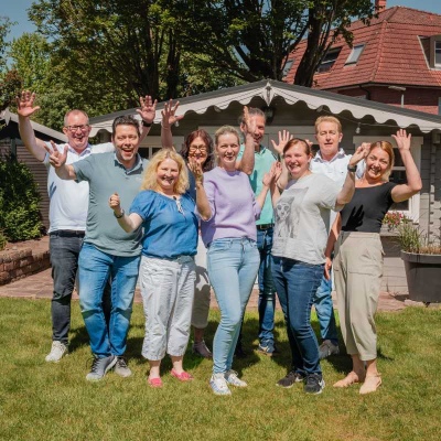 Das Team der Steuerkanzlei Möller steht draußen vor einem Haus, lächelt und winkt in die Kamera. Es ist sonnig, alle wirken fröhlich und entspannt.